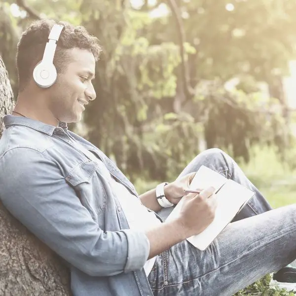 Guy resting on tree listening to music and writing down his goals